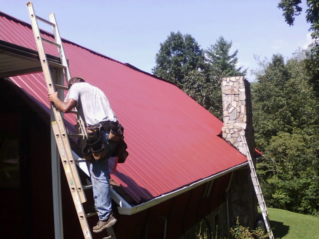 Roofer on a ladder beside a red metal roof and stone chimney — Soffit & Fascia Repair by Roofing Brothers in Hackensack